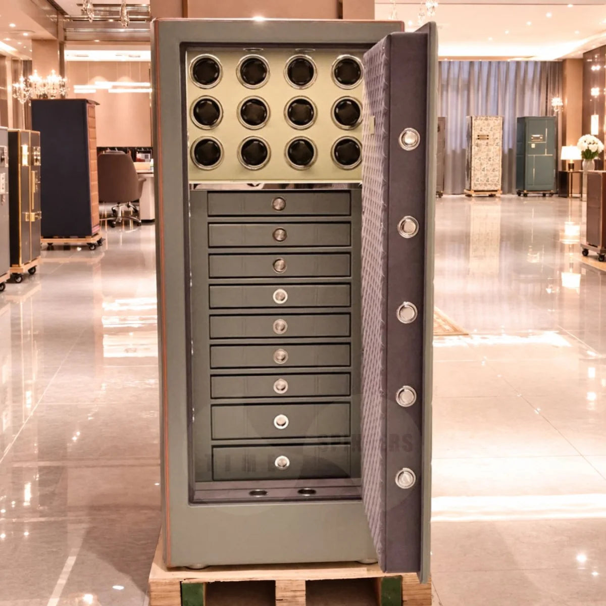 A large gray metal safe cabinet with an open door, showing a top section with circular watch winder holders and a lower section with multiple drawers, photographed in a polished showroom interior.